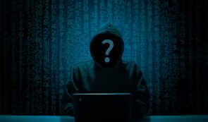 A hooded figure with a question mark symbol on their face working on a laptop against a background of falling digital code.