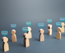Wooden figurines with speech bubble icons above their heads, representing group conversation or communication on a gray background.