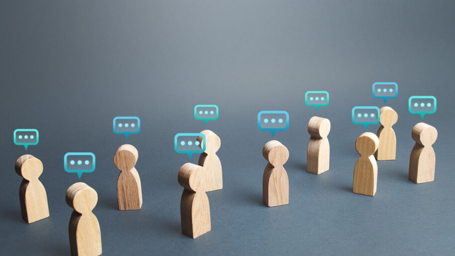 Wooden figurines with speech bubble icons above their heads, representing group conversation or communication on a gray background.