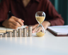 Rising coin stacks beside an hourglass on a desk, representing long-term investment growth over time.