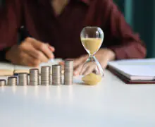 Rising coin stacks beside an hourglass on a desk, representing long-term investment growth over time.