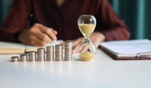 Rising coin stacks beside an hourglass on a desk, representing long-term investment growth over time.
