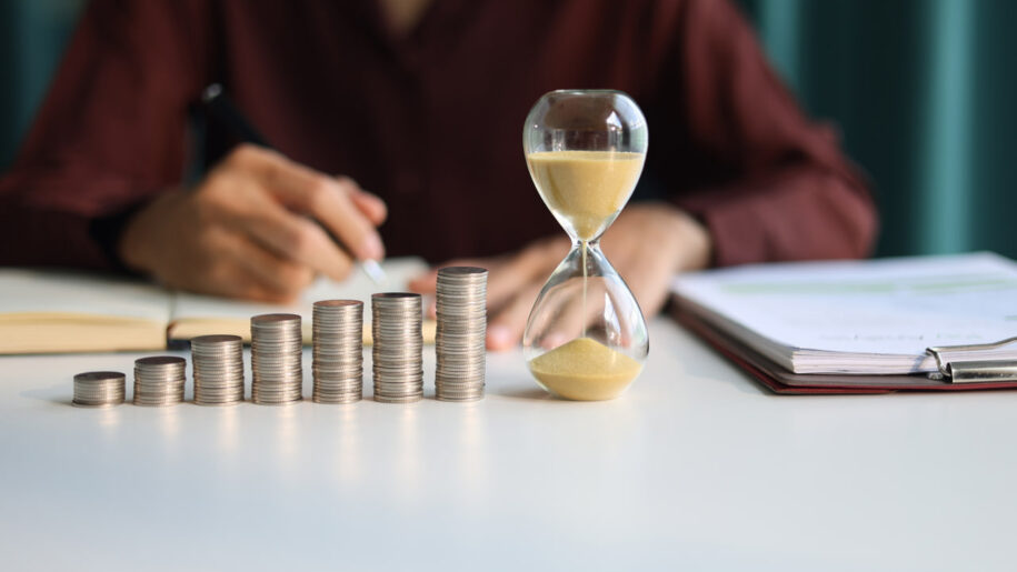 Rising coin stacks beside an hourglass on a desk, representing long-term investment growth over time.