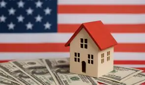 A small model house resting on top of US dollar bills with an American flag background.