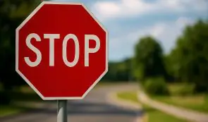 Red octagonal stop sign posted on the side of a paved road with trees and a sidewalk in the background.
