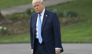 Donald Trump walking outside in a dark blue suit and light blue tie against a green lawn background.