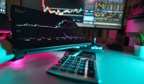 A glowing trading desk setup featuring a tablet displaying candlestick charts, a calculator, and multiple monitors showing market data and technical indicators in a neon-lit room.