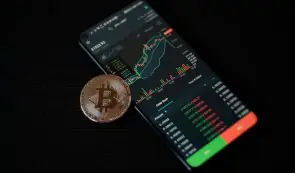 Smartphone displaying a crypto trading application with candlestick charts alongside a physical Bitcoin token on a dark surface.