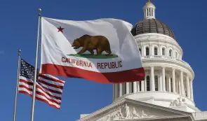 California State Capitol building in Sacramento with the California Republic bear flag and U.S. flag flying in the foreground against a blue sky.