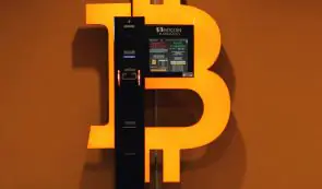Bitcoin ATM machine mounted against an illuminated orange Bitcoin logo wall.