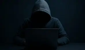Anonymous figure in a hoodie working on a laptop in the dark.