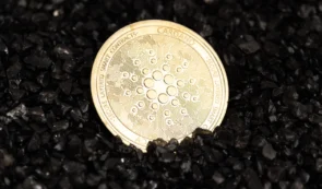 Physical silver Cardano (ADA) cryptocurrency coin resting on a surface covered in dark black gravel.