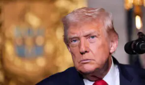 Donald Trump speaking at a press conference against a blurred gold backdrop, relevant to digital asset policies and institutional crypto staking frameworks.