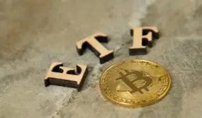 Wooden letters spelling "ETF" alongside a physical gold Bitcoin token resting on a textured surface.