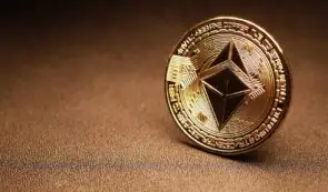 A physical gold Ethereum coin resting on a textured surface.