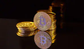 Stacks of gold Bitcoin physical tokens reflecting on a dark, reflective surface against a dark background.