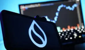 Smartphone displaying the Sui cryptocurrency logo with financial charts on a monitor in the background.