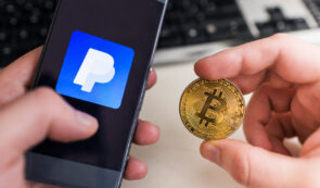 Two hands holding a smartphone displaying the PayPal logo and a physical Bitcoin coin, with a keyboard visible in the background