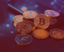 Group of physical cryptocurrency coins including Bitcoin, Ethereum, Ripple, Dogecoin, and Cardano, resting on a smartphone.