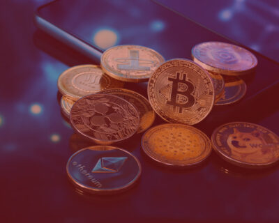 Group of physical cryptocurrency coins including Bitcoin, Ethereum, Ripple, Dogecoin, and Cardano, resting on a smartphone.