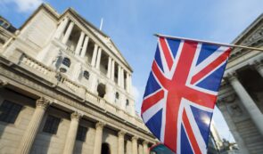 A handheld UK Union Jack flag in the foreground with the grand neoclassical facade of the Bank of England building in London in the background under a blue sky.