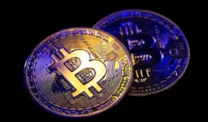 Two physical Bitcoin coins illuminated with blue and gold lighting against a black background.
