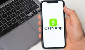 A hand holding a smartphone displaying the green Cash App logo and wordmark on screen with a laptop keyboard in the background.
