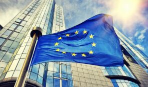 European Union flag waving in front of a modern glass and steel European institution building.