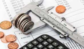 A caliper measuring a stack of coins resting on financial documents alongside a calculator and loose coins.
