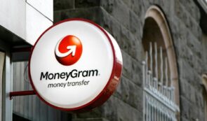 Round illuminated MoneyGram money transfer sign mounted on a building wall with stone architecture in the background.