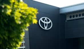 The silver Toyota logo mounted on the dark exterior wall of a modern corporate dealership building next to green foliage.