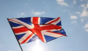 The Union Jack flag of the United Kingdom waving proudly against a bright blue sky with scattered clouds.