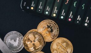 Alt: Physical cryptocurrency tokens including Bitcoin, XRP, and Litecoin arranged next to a smartphone displaying a crypto exchange market app interface.