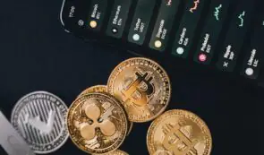 Alt: Physical cryptocurrency tokens including Bitcoin, XRP, and Litecoin arranged next to a smartphone displaying a crypto exchange market app interface.