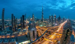 Aerial evening view of Dubai skyline illuminated at dusk, featuring the Burj Khalifa and glowing highway traffic trails.