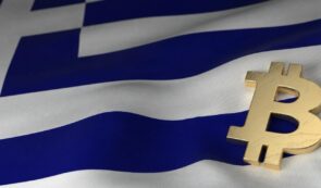 A golden physical Bitcoin symbol resting against the backdrop of the Greek national flag.