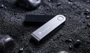 A Ledger hardware cryptocurrency wallet resting on a textured, dark gravel surface.
