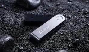 A Ledger hardware cryptocurrency wallet resting on a textured, dark gravel surface.