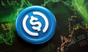 Physical blue and white USD Coin (USDC) stablecoin token placed over a background displaying glowing financial market trading charts and data.