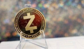 Physical Zcash (ZEC) cryptocurrency token with a gold-colored finish and the Z symbol displayed on a clear display stand against a sparkling bokeh background.
